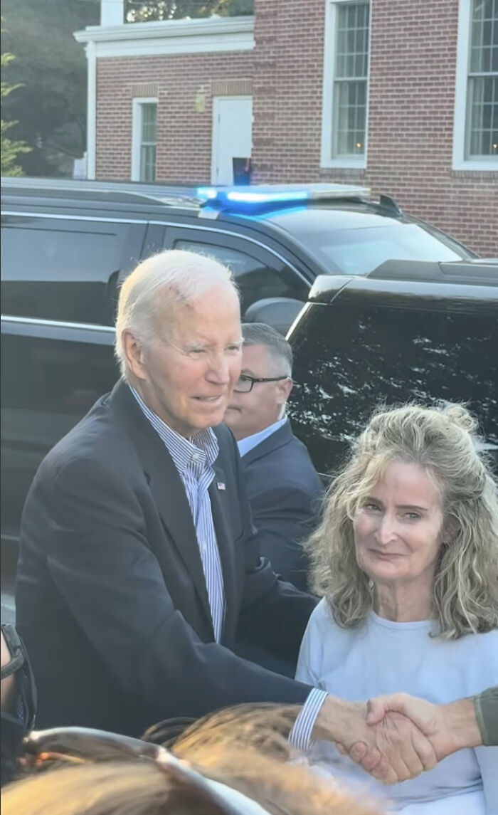 Joe Biden with a visible head wound, shaking hands with a woman outside near black vehicles and a brick building.