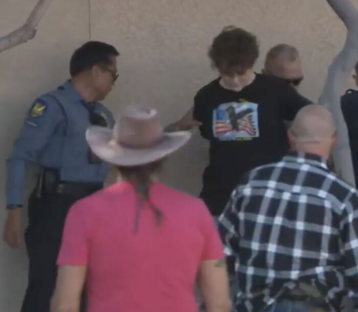 Teen wearing shirt like accused Charlie Kirk shooter, surrounded by police and others at a memorial event.