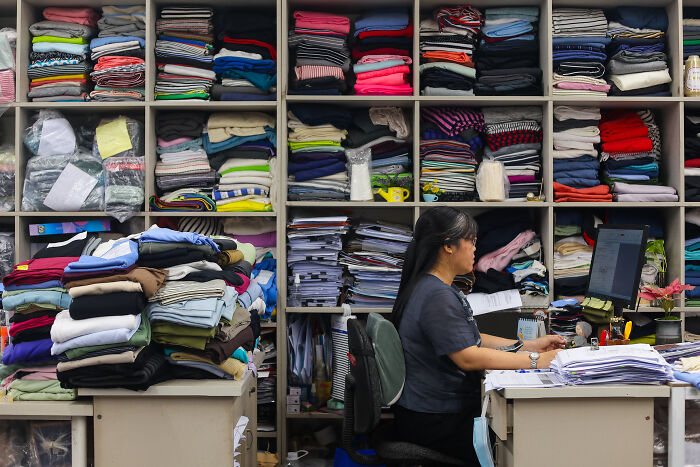 Woman working in a clothing warehouse surrounded by stacks of folded clothes, illustrating Trumps trade war costing billions impact.