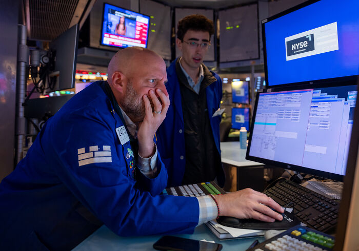 Two traders in blue jackets at the NYSE, focused on screens showing market data amid trade war costing billions concerns.