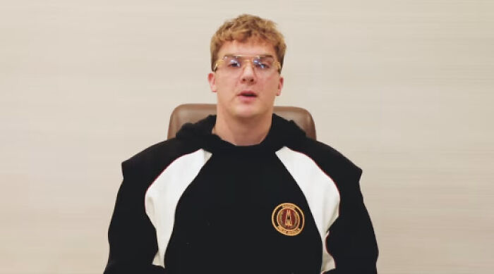 Young man in glasses and black hoodie speaking in a plain setting about creators of Melania Trump meme coin crypto fraud.