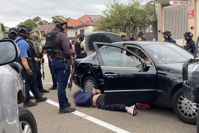 Armed law enforcement intercepts alleged assassins near vehicle in a public parking lot during a staged operation.