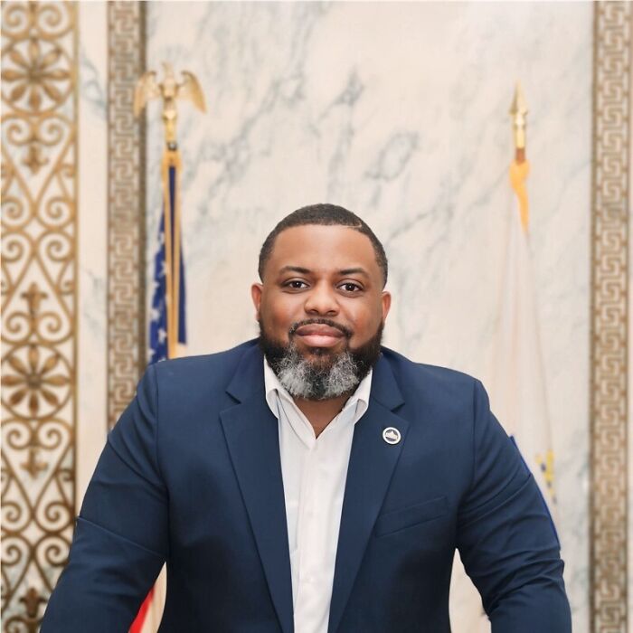 Man in navy suit with white shirt posing in an official government office setting with flags in the background.