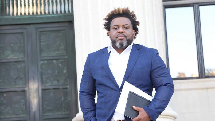 Man in a blue suit standing outside a government building, holding documents related to aide arrest for c*****e shipment.