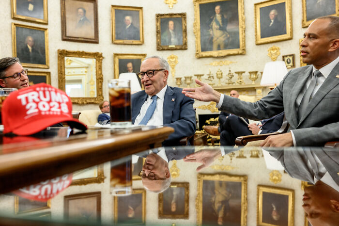 Politicians in a formal meeting room, engaged in a discussion amid government shutdown and partisan blame efforts.