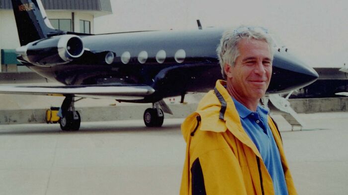 Jeffrey Epstein standing on an airport tarmac near a private jet, wearing a yellow jacket and blue shirt.