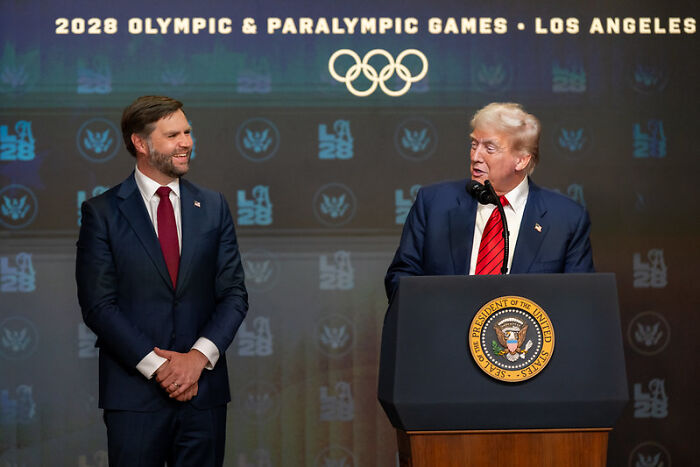 JD Vance in a suit standing near a podium with a man speaking, backdrop showing 2028 Olympic and Paralympic Games logos.