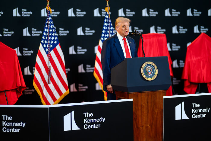 Former President Trump speaking at The Kennedy Center podium with American flags in the background on a formal stage setting.