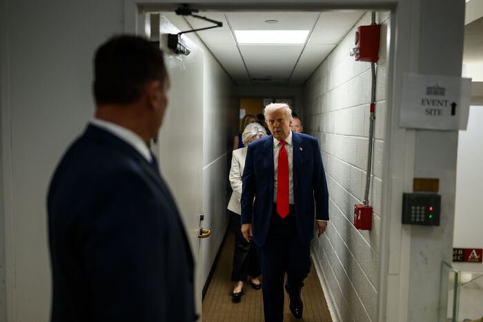 Man in a suit and red tie walking down a narrow corridor, with others following behind, related to ICE deportation incident.