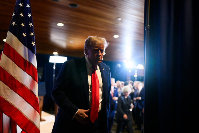 Donald Trump standing near an American flag backstage with bright lights, linked to Bernie Sanders sombrero troll campaign.