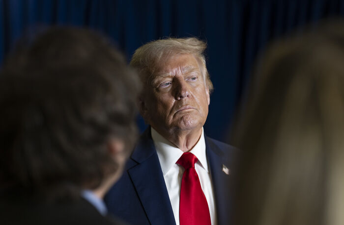 Former President Trump in a dark suit and red tie during a media event, highlighting government focus instead of American people.
