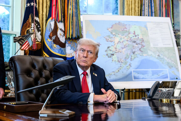Donald Trump sitting at the desk in the Oval Office with a map of Alaska in the background during a formal meeting.