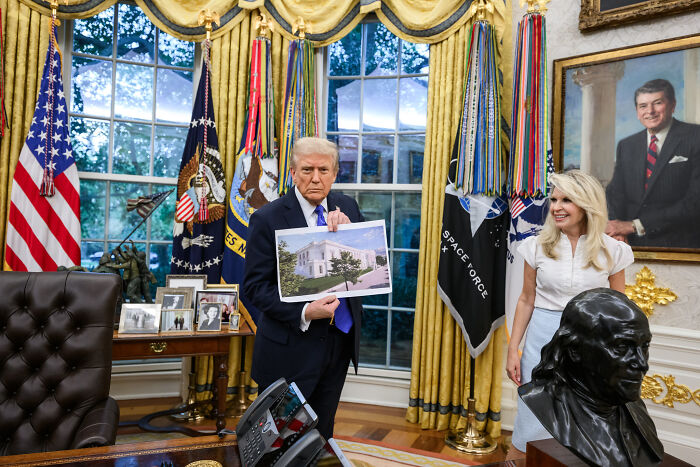 Donald Trump in the Oval Office holding a photo, related to agency oversight of controversial D.C. renovations.