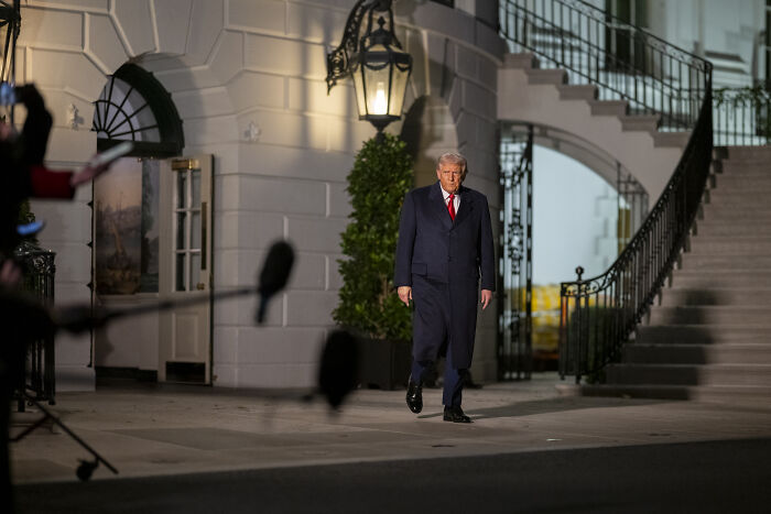 Former President Trump walking outside White House at night during media coverage on visa-related issue.