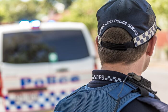 Police officer from Queensland Police Service facing away, standing near a police van at an outdoor scene.