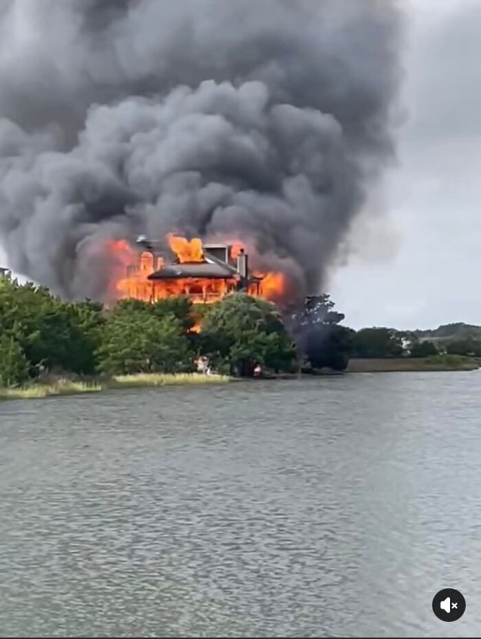 House of judge engulfed in flames with thick black smoke rising, surrounded by trees near a body of water.