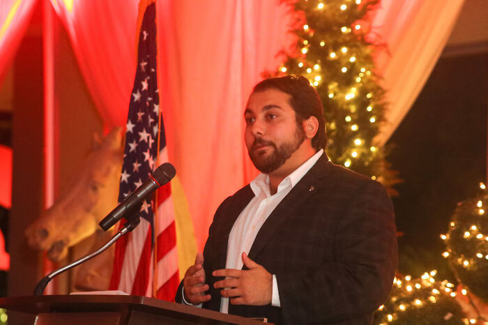 Young Republican leader speaking at a podium with an American flag, amid holiday lights and decorations.