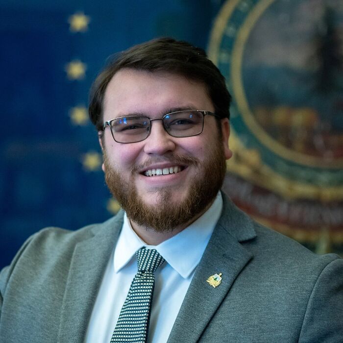 Young Republican leader wearing glasses and suit smiling in an official setting with political emblem background.