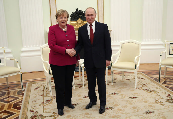 Angela Merkel shaking hands with Vladimir Putin in a formal room, related to Poland and Baltic States fueling Putin&rsquo;s war.