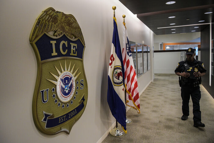 ICE emblem on wall with US and agency flags, a uniformed officer walking down a hallway inside a government facility.