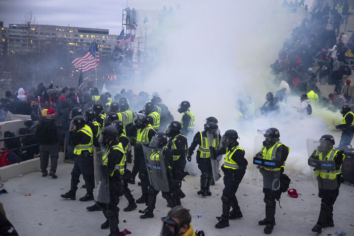 Police in riot gear standing amid smoke during January 6 event with crowd in background, highlighting prosecutors and DOJ leave.
