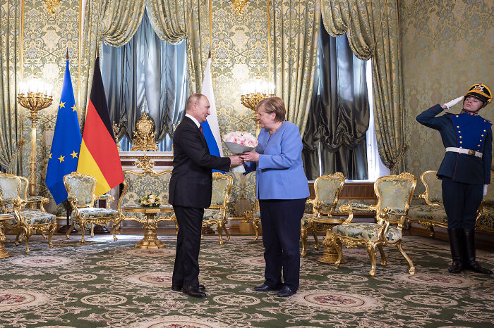 Angela Merkel and Vladimir Putin exchanging flowers in an ornate room with European and German flags.