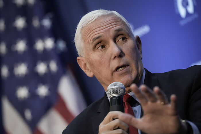 Mike Pence speaking at an event, holding a microphone, with a blurred American flag background, related to Trump called Pence wimp.