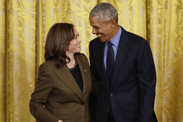 Barack Obama and Kamala Harris smiling at each other in a formal setting with gold curtains in the background