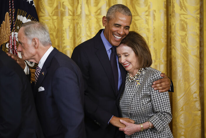 Barack Obama warmly hugging Nancy Pelosi in a formal setting with golden curtains in the background.