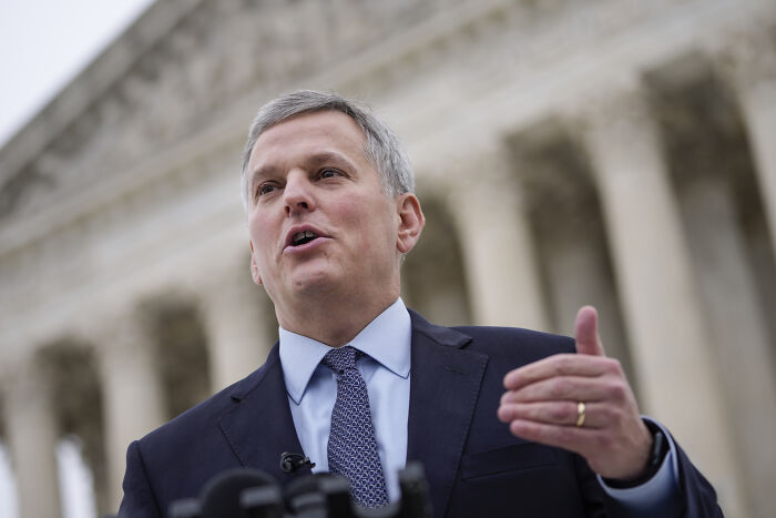 Man in suit speaking at an outdoor podium with columns in background, referencing Ukrainian refugee assailant law.