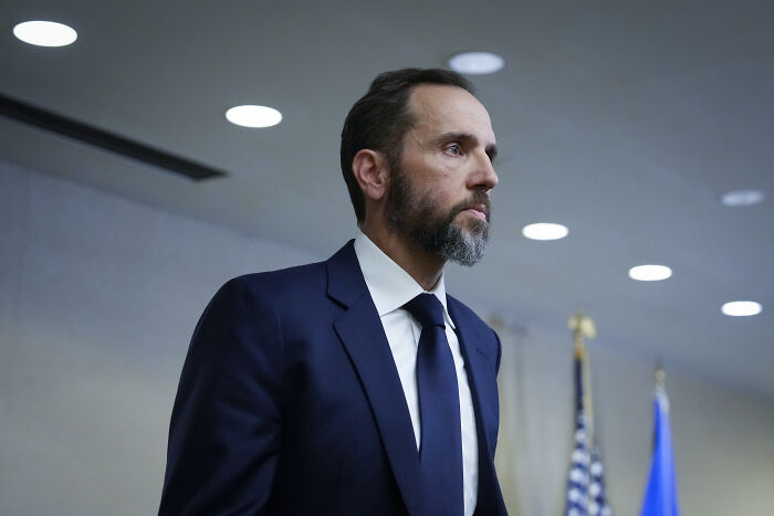 Man in a dark suit and tie with a beard, standing indoors near American flags during a January 6 investigation event.