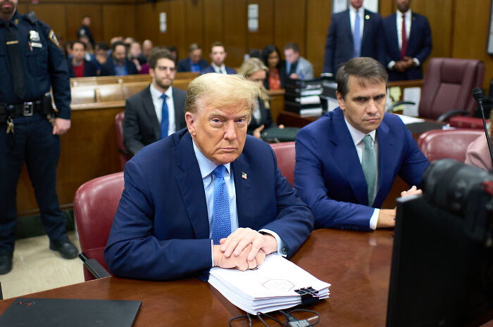 Trump at a courtroom table with legal documents, defending $230 million compensation bid against DOJ.