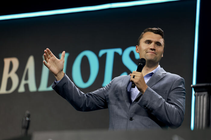 Man in gray suit speaking into microphone on stage with partial ballot text in background, related to Charlie Kirk protest.