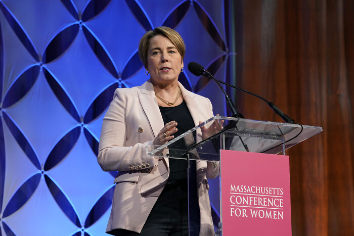 Aide to Gov Maura Healey speaking at Massachusetts Conference for Women event, wearing a light blazer at a clear podium.