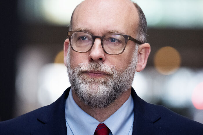 Russ Vought wearing glasses and a suit, known for Project 2025, pictured discussing agency budget cuts.
