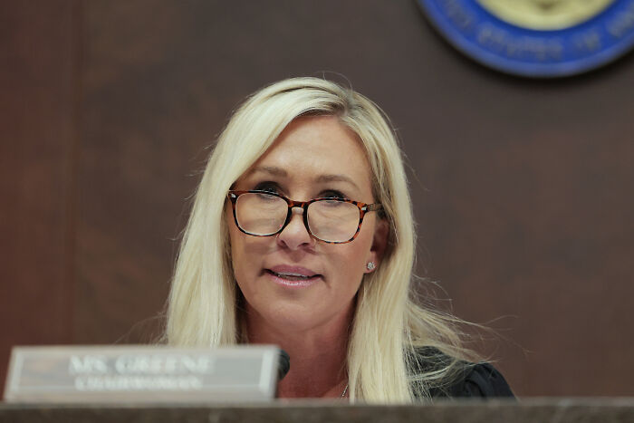 Marjorie Taylor Greene speaking in a formal setting, wearing glasses and seated before a microphone.