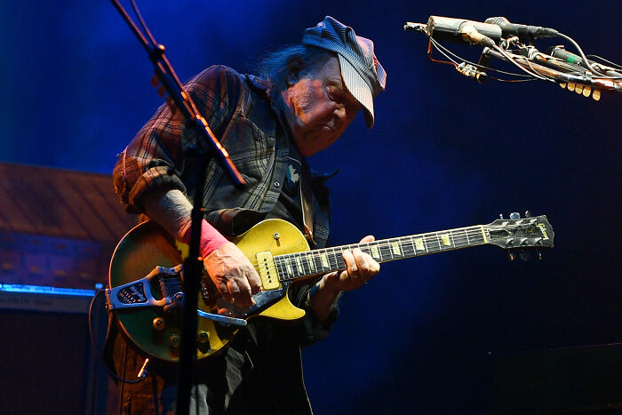 Neil Young playing electric guitar on stage, highlighting protest against Trump-aligned corporations and Amazon music removal.