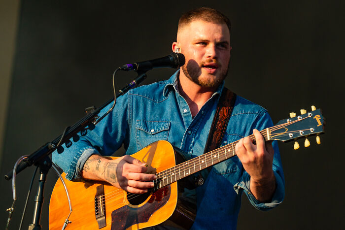 Country musician performing on stage with acoustic guitar and microphone, related to anti-ICE song controversy.