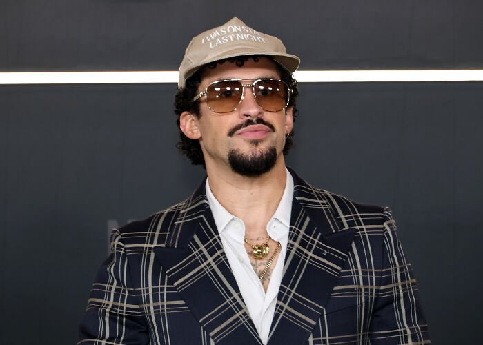 Bad Bunny wearing sunglasses and a hat, posing in a plaid blazer ahead of Super Bowl performance with ICE agents mentioned.