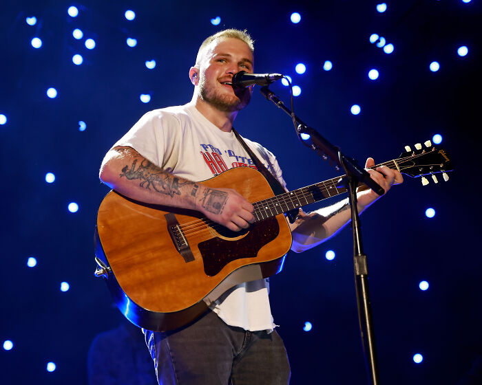 Country musician performing on stage with an acoustic guitar under blue lights relating to anti ICE song controversy.