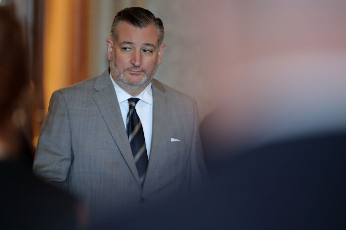 Ted Cruz in a gray suit and striped tie, looking to the side during a formal event with blurred people in foreground.