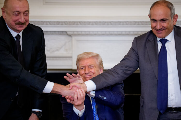 Donald Trump smiling between two men shaking hands at European summit amid geographic gaffe mockery by leaders