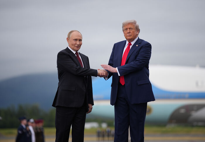 Donald Trump and Vladimir Putin shaking hands outdoors with airplane in background, related to Trump Putin meeting and nuclear tension.