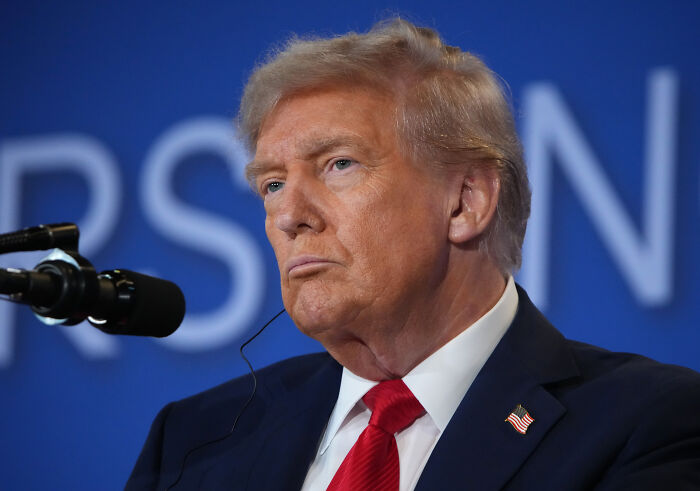 Donald Trump speaking at a microphone, dressed in suit and red tie, with blue background featuring blurred text.