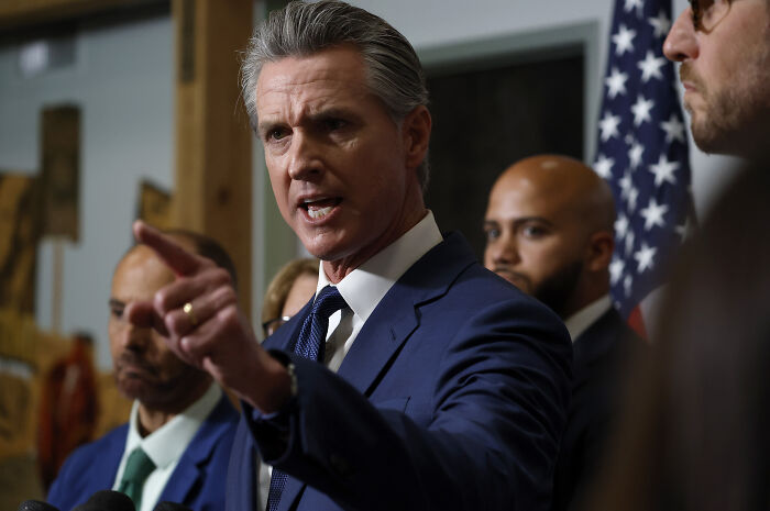 Man in blue suit pointing and speaking at a podium with others behind him and an American flag in the background