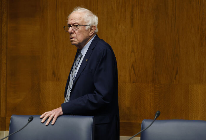 Bernie Sanders standing in a navy suit and striped tie in a wood-paneled room during a formal event.
