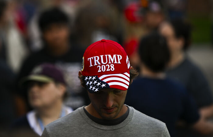 Man wearing red Trump 2028 hat with American flag design, representing hints of presidency continuing indefinitely.