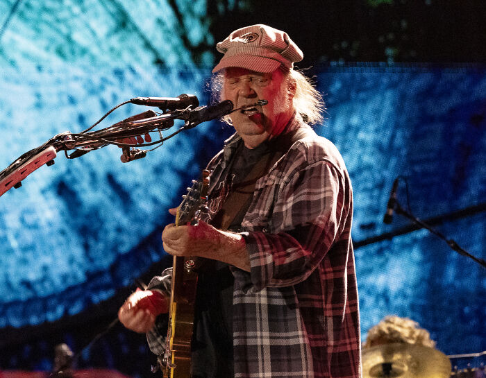 Neil Young performing live on stage with guitar and harmonica during a music protest against Amazon and Trump-aligned corporations.