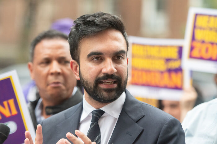 Man in a suit speaking passionately at an outdoor event, with placards in the background about political campaigns.