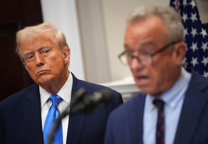 Donald Trump listening intently while a man in glasses speaks at a formal event with an American flag visible.